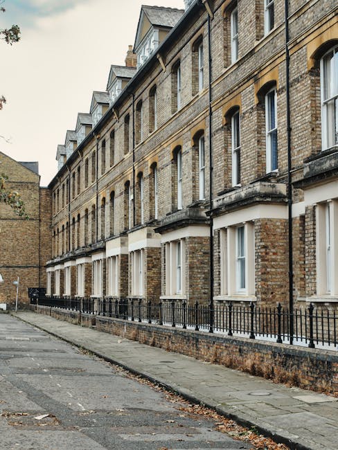 A narrow street in a residential area featuring a row of traditional brick terraced houses with large, multi-pane sash windows and small front gardens bordered by black metal railings. The sidewalk is