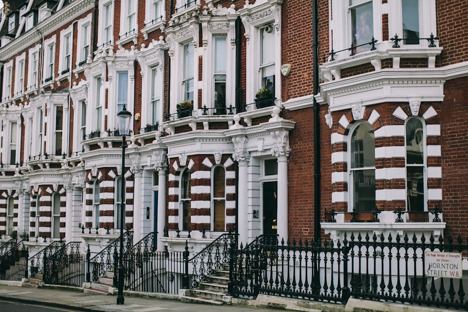 A narrow street in a residential area featuring a row of traditional brick terraced houses with large, multi-pane sash windows and small front gardens bordered by black metal railings. The sidewalk is