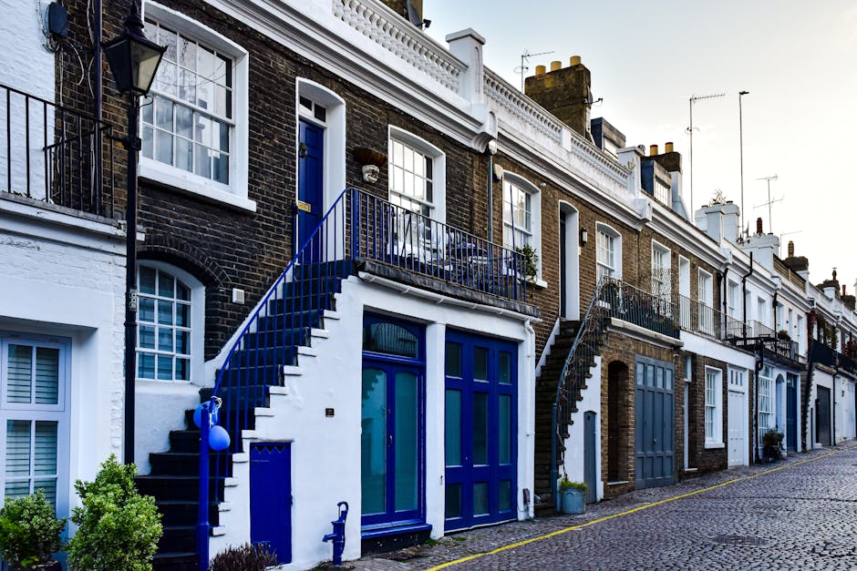 A row of terraced residential buildings on New Kings Road Fulham, with exterior staircases in black and blue leading to first-floor entrances, and ground-floor glass doors and windows. The houses feature a mix of brick and painted facades, with some showing white plaster and others exposed brickwork. Each property has a small balcony or terrace with metal railings, and the street is paved with cobblestones. A few potted plants are visible near doorways, and a blue plastic container is placed beside one of the staircases. The scene is outdoors, captured during daylight, illustrating an urban environment suitable for house removals and furniture transport, with Removals Companies Fulham occasionally involved in the moving process depicted in similar settings.