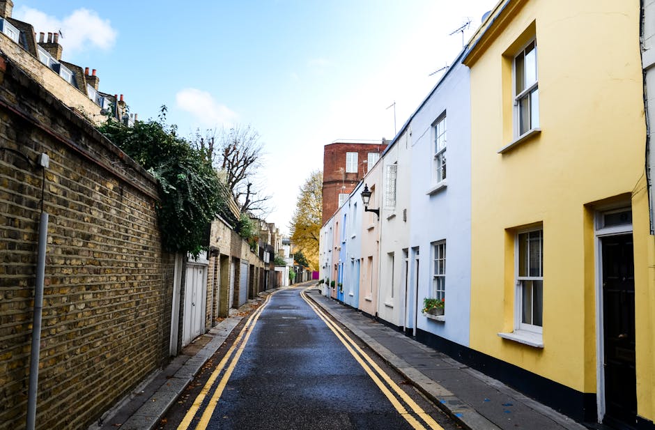 A row of terraced residential buildings on New Kings Road Fulham, with exterior staircases in black and blue leading to first-floor entrances, and ground-floor glass doors and windows. The houses feature a mix of brick and painted facades, with some showing white plaster and others exposed brickwork. Each property has a small balcony or terrace with metal railings, and the street is paved with cobblestones. A few potted plants are visible near doorways, and a blue plastic container is placed beside one of the staircases. The scene is outdoors, captured during daylight, illustrating an urban environment suitable for house removals and furniture transport, with Removals Companies Fulham occasionally involved in the moving process depicted in similar settings.