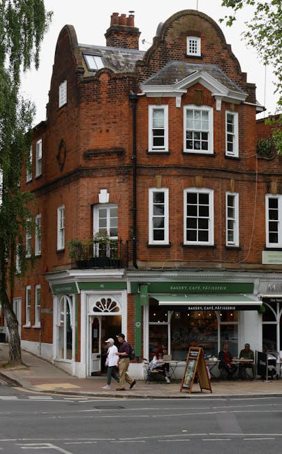 A three-story red brick building with a curved gabled roof and decorative white trim, situated on a street corner. The ground floor features a bakery, café, and pâtisserie with large windows and a green awning displaying the names. Outside, several people are walking past or sitting at outdoor tables on the pavement. The scene includes an assortment of cardboard boxes and packing materials placed near the entrance, indicating potential house removal or moving preparations, with a green and black trolley possibly used for loading. The surrounding environment includes leafy trees, a paved sidewalk, and modern street infrastructure. The presence of packed boxes and the activity of people suggest ongoing furniture transport or relocation efforts, which [COMPANY_NAME] can assist with, especially for efficient moving logistics in Fulham.