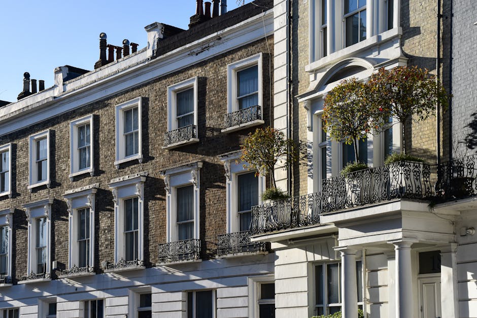 The image depicts a brick residential building on Melbourne Terrace in Fulham, London, with three white-framed windows visible on the upper floor, two of which are open. The ground floor features two doorways with dark doors, one of which is partially open, revealing a glimpse of the interior, and each doorway is topped with a brick arch. To the left of the doors, there is a small wall-mounted light fixture. Adjacent to the building, a street sign indicates the location as Moore Park Road, SW6, and another sign warns about road humps for 450 yards. The scene suggests an urban environment suitable for house removals or home relocation projects, with potential logistics involving moving furniture, packing materials, and loading activities occurring outside the property.