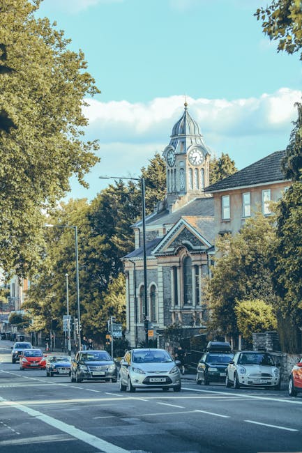 A row of parked cars along a street in Fulham, with various models including hatchbacks and sedans, positioned on the street pavement next to a sidewalk with a few trees providing shade. In the background, a historic-looking church with a prominent clock tower topped with a spire is visible, featuring arched windows and detailed architectural features. The scene is set on a clear day with blue sky and some white clouds, and the street appears to be in a residential or mixed-use area. The photograph, taken during daylight hours, captures the exterior environment where furniture and belongings could be loaded during a home relocation. The presence of parked vehicles and the urban setting aligns with typical moving logistics and packing activities, with potential involvement of professional removal services like Removal Companies Fulham.