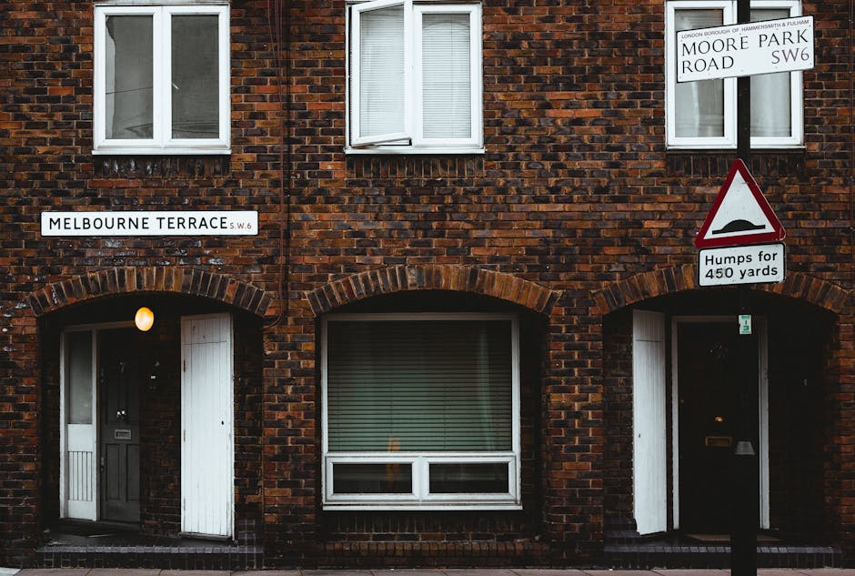 Photograph of a brick residential building on Melbourne Terrace in Fulham, displaying two ground-floor doorways with white wooden doors, one slightly open, and a large rectangular window with closed blinds between them. Above each doorway is an archway made of red bricks, and a single exterior light is mounted next to the left door. Adjacent to the right doorway, a white street sign indicates the location as Moore Park Road SW6. A triangular warning sign with a hump icon and the message 'Humps for 450 yards' is mounted on a pole in front of the building. The façade features brickwork with a mix of dark and reddish tones, and there are white-framed windows and a sign reading 'MELBOURNE TERRACE SW6' on the wall. The street scene suggests a typical residential area where house removals and furniture transport could take place, with natural daylight illuminating the scene, providing context for moving or packing activities associated with home relocation services by Removal Companies Fulham.
