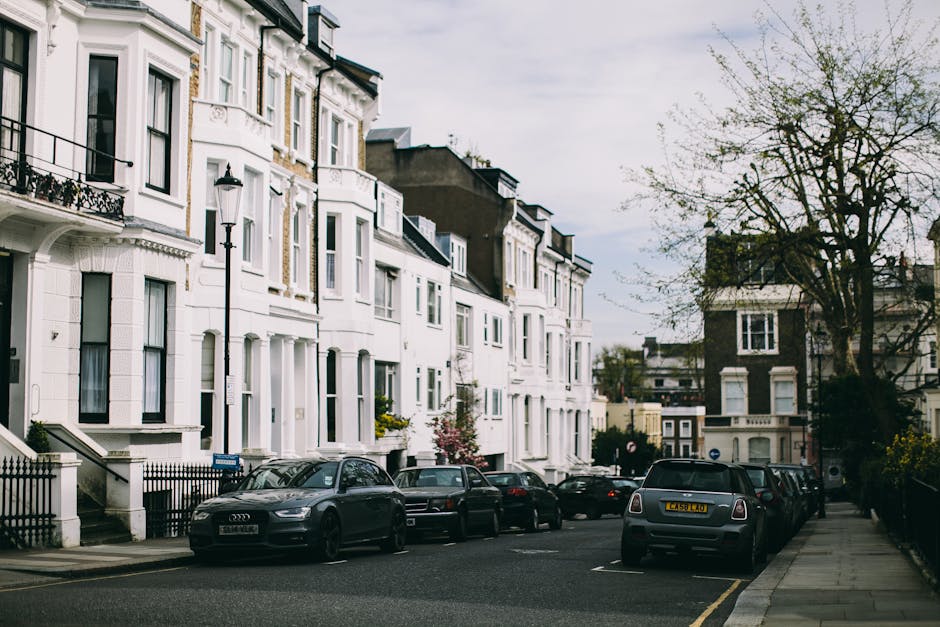 A row of parked cars along a street in Fulham, with various models including hatchbacks and sedans, positioned on the street pavement next to a sidewalk with a few trees providing shade. In the background, a historic-looking church with a prominent clock tower topped with a spire is visible, featuring arched windows and detailed architectural features. The scene is set on a clear day with blue sky and some white clouds, and the street appears to be in a residential or mixed-use area. The photograph, taken during daylight hours, captures the exterior environment where furniture and belongings could be loaded during a home relocation. The presence of parked vehicles and the urban setting aligns with typical moving logistics and packing activities, with potential involvement of professional removal services like Removal Companies Fulham.